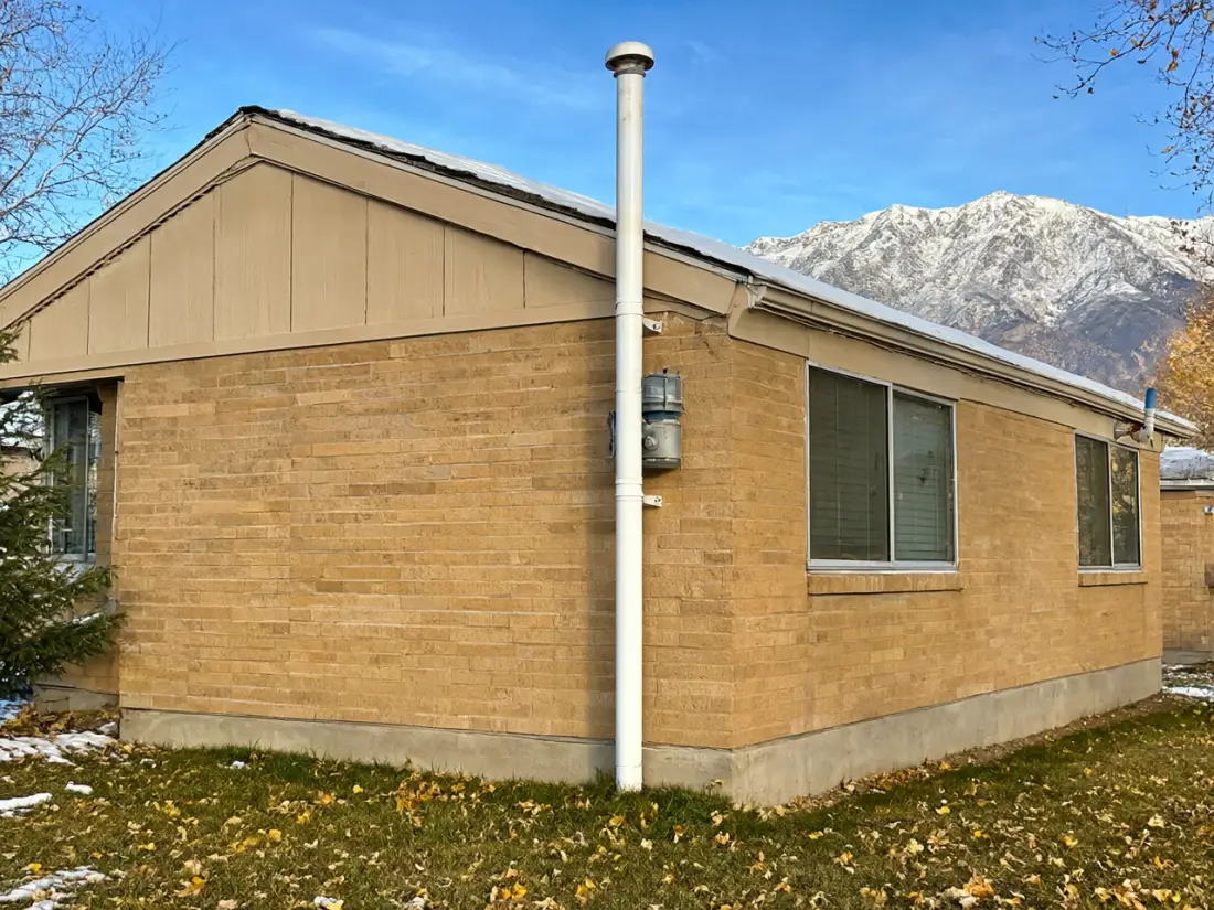 A white radon vent pipe running up the side of a brick house to above the roofline, with the fan mounted on the outside wall. Placeholder image.
